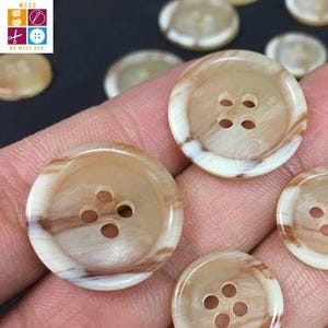 May include: Close-up of several round buttons with four holes each. The buttons have a marbled appearance with shades of tan, brown, and cream. The buttons are being held in a hand. The logo "MISS BY MISS DEY" is in the upper left corner.