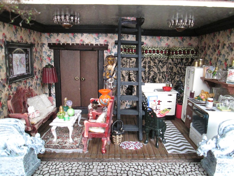 May include: A miniature dollhouse interior with a living room and kitchen. The living room has a floral wallpaper, a red velvet couch, a coffee table, and a lamp. The kitchen has a white refrigerator, a black stove, and a wooden countertop. There is a ladder leading to an upper level.