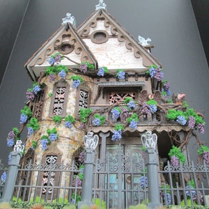 May include: A miniature dollhouse with a gothic design, featuring a white facade with brown trim, a black wrought iron fence, and a lush green ivy vine with purple flowers.