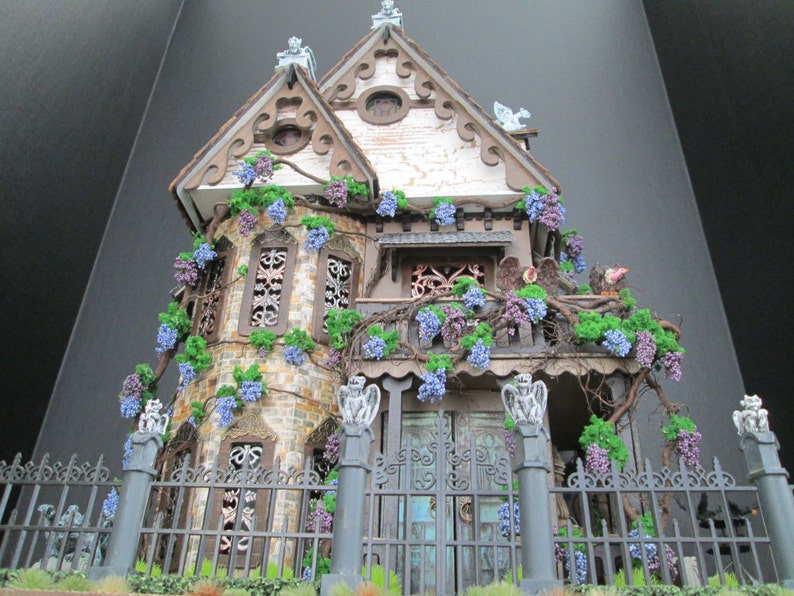 May include: A miniature dollhouse with a gothic design, featuring a white facade with brown trim, a black wrought iron fence, and a lush green ivy vine with purple flowers.