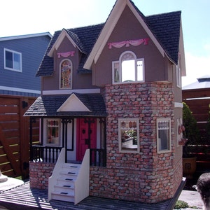May include: A detailed dollhouse with a brown exterior, brick accents, and a black shingled roof. The house features a pink front door, white trim, and decorative pink ribbons. The dollhouse has multiple windows and a small porch with a railing.
