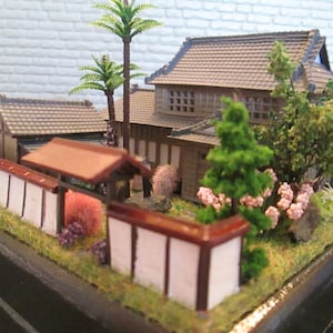May include: A miniature Japanese garden scene with a small house, a fence, and various trees and plants. The house has a brown roof and white walls, and the fence is made of wood and has a red top. The trees and plants are green and brown, and there are some pink flowers in the garden.