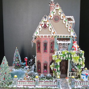 May include: A miniature Christmas house with a red brick facade, white trim, and a snow-covered roof. The house has a porch with a green garland and a snowman standing in front. There are two Christmas trees in the yard, one with red and green ornaments and the other with white lights.