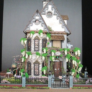 May include: A miniature dollhouse with a white exterior, gray stone accents, and a green roof. The house has a front porch with a wooden door and a green wreath. The house is surrounded by a black wrought iron fence and a lush green lawn.