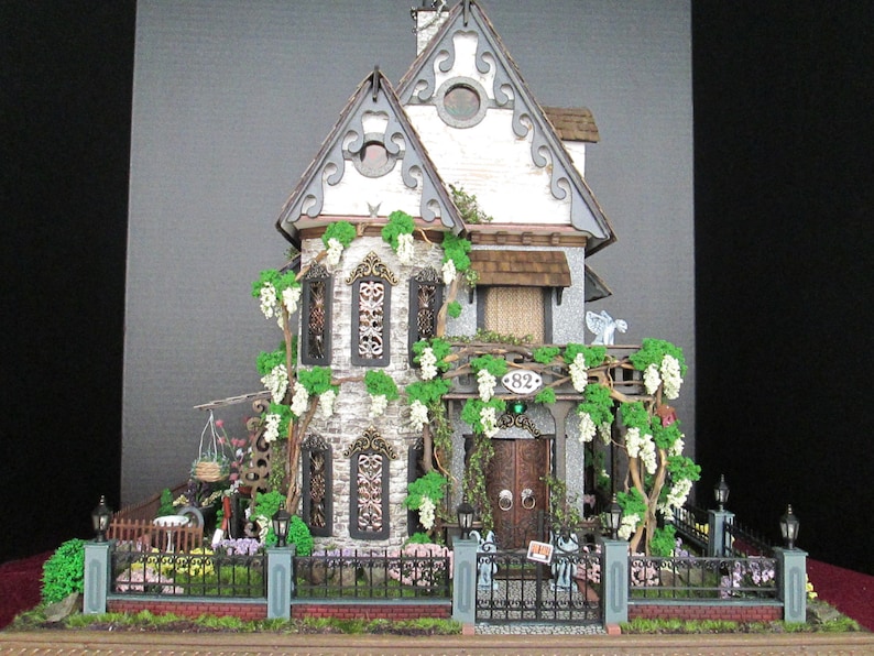 May include: A miniature dollhouse with a white exterior, gray stone accents, and a green roof. The house has a front porch with a wooden door and a green wreath. The house is surrounded by a black wrought iron fence and a lush green lawn.