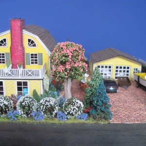 May include: A miniature model of a yellow house with a red chimney and a white porch, surrounded by lush green bushes and trees. A smaller yellow garage is in the background with a black car parked in front of it. A small white boat is parked on the driveway.