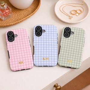 May include: Three phone cases in pink, blue, and green, each with a gingham pattern. The cases have black edges and gold initials. A small dish with jewelry and a perfume bottle are in the background.