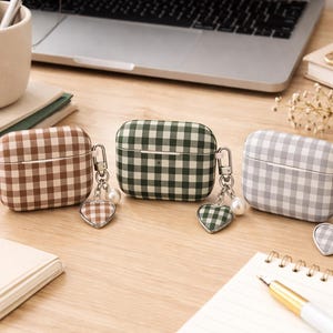 May include: Three AirPod cases in brown, green, and gray gingham patterns. Each case has a silver-tone clasp and a heart-shaped charm with a pearl accent. The cases are on a wooden surface with office supplies and a laptop in the background.
