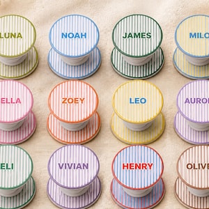 May include: A collection of personalized phone grips with names like Luna, Noah, and James. Each grip features a striped design in various colors, with the name printed in a matching hue. The grips are round and have a collapsible design.