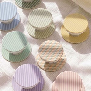 May include: A collection of eight circular phone grips featuring various pastel striped patterns in shades of green, blue, yellow, purple, and pink. Each grip has a flat, round top surface with vertical stripes and a matching circular base.