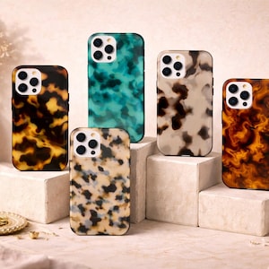 May include: A collection of five phone cases with unique patterns. The cases feature designs in shades of brown, teal, and cream, resembling tortoiseshell and abstract textures. They are displayed on small, square pedestals, with a candle and decorative items in the background.