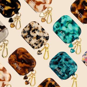 May include: A collection of personalized AirPod cases in various colors and patterns, including tortoiseshell, marble, and teal. Each case features a gold-colored keychain with heart and gem charms, and some have names or initials.