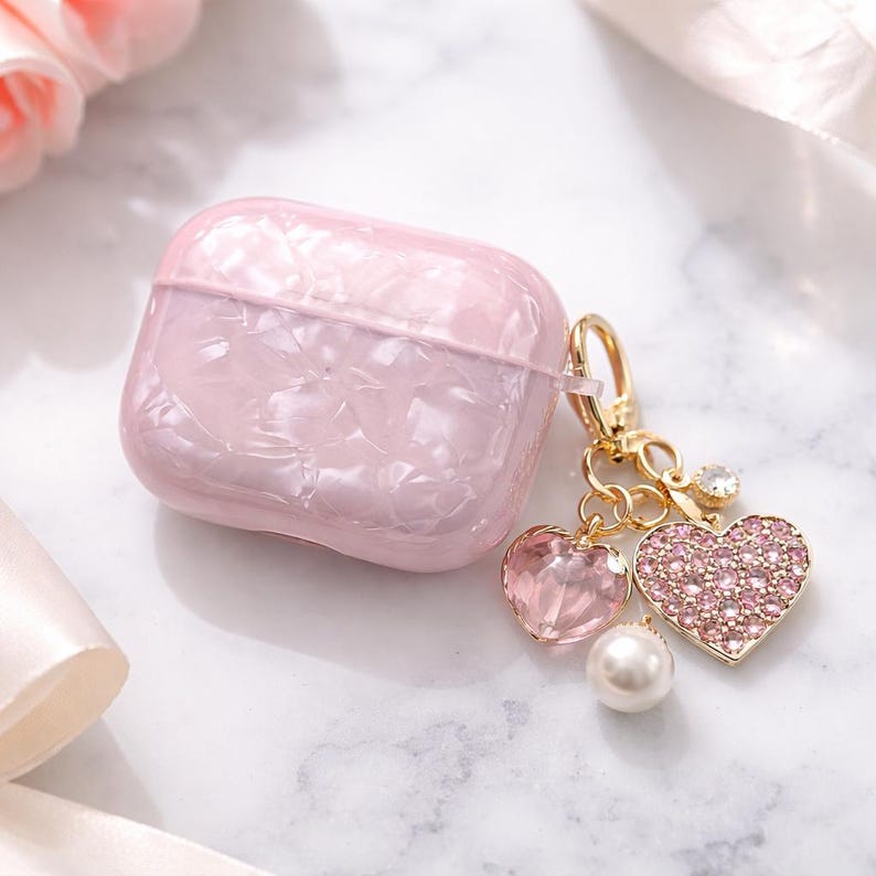May include: A pink marbled AirPod case with a gold-tone charm. The charm has a clear heart-shaped gem, a pink rhinestone-covered heart, and a white pearl. The case is square with rounded edges.