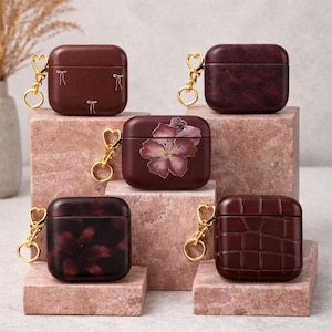 May include: Five maroon-colored AirPod cases with gold-colored heart-shaped keychains. The cases have different designs, including a floral print, a textured pattern, and a bow design. The cases are displayed on pink blocks.