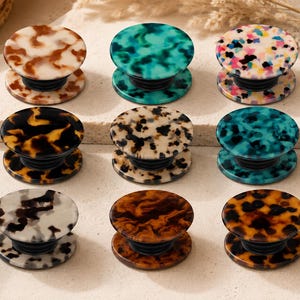 May include: A collection of nine decorative phone grips in various colors and patterns. The grips feature a round top and base with a collapsible design. Colors include tortoiseshell, teal, and speckled patterns. The grips are displayed on a neutral surface.