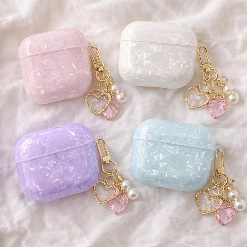 May include: Four iridescent AirPod cases in pastel shades: pink, white, lilac, and blue. Each case features a gold-coloured keychain with heart charms and faux pearls. The cases have a geometric, textured pattern.