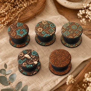 May include: Five black phone grips with decorative tops. The tops feature floral and geometric patterns in shades of brown, teal, and orange. One grip has a skull and floral design. The grips are on a tan surface.