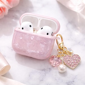 May include: A pink marble-effect AirPod case containing white earbuds. The case has a gold-coloured clasp and a charm with a pink gem heart, a pearl, and a rhinestone heart. The case is on a white marble surface.