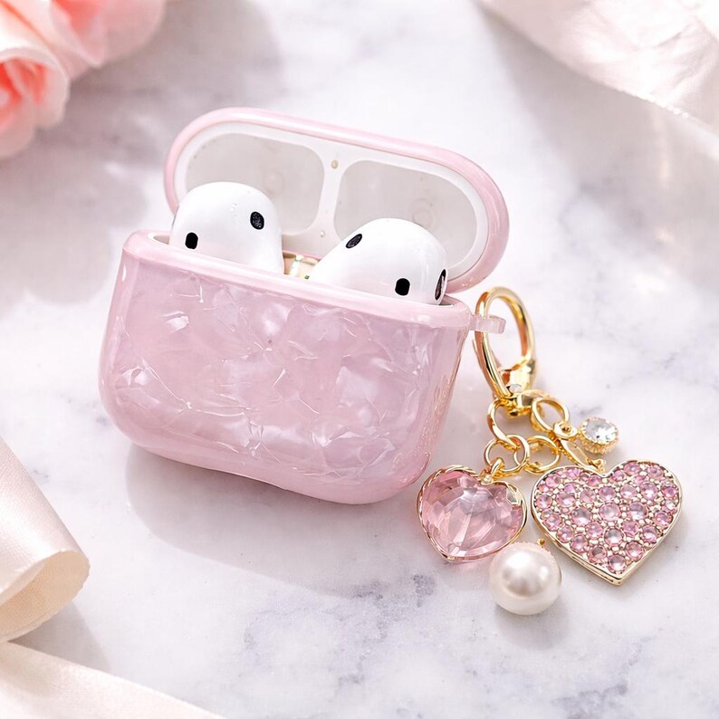 May include: A pink marble-effect AirPod case containing white earbuds. The case has a gold-coloured clasp and a charm with a pink gem heart, a pearl, and a rhinestone heart. The case is on a white marble surface.