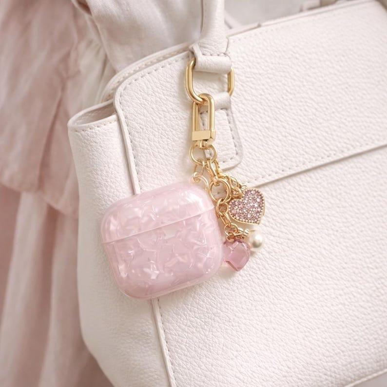 May include: A pink, patterned AirPod case with a gold-tone keychain and heart charms. The case is attached to a white handbag. The charms include a rhinestone-covered heart, a pearl, and a small pink heart.