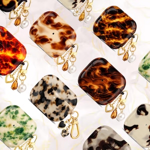May include: A collection of AirPod cases with various patterns, including tortoiseshell, speckled, and floral designs. Each case features a gold-colored charm with heart and pearl accents, and a gold-colored clasp.