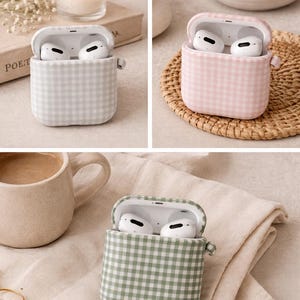 May include: Three AirPods cases in different colors: gray, pink, and green. Each case has a gingham pattern and is open, revealing white earbuds. The cases are displayed with a candle, a cup, and decorative items.