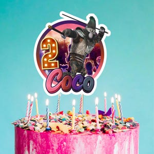 May include: A pink frosted birthday cake with colorful sprinkles and lit candles. A cake topper features a cartoon gorilla character with the number "2" and the name "Coco". The background is a light blue.