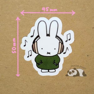 May include: A white bunny sticker wearing headphones and a green dress, with musical notes around it. The sticker is 50mm tall and 45mm wide. A small panda illustration and the text "Zanda Studios" are also present.