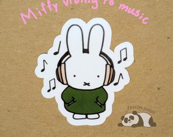 May include: A white cartoon bunny wearing headphones and a green dress, surrounded by musical notes. The text "Miffy vibing to music" is written above the bunny. A sleeping panda is in the lower right corner. The sticker is from Zanda Studios.