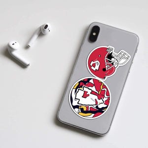 May include: A smartphone with two stickers featuring a red football helmet and a circular design with red, yellow, and white elements. White wireless earbuds are placed on the white surface.