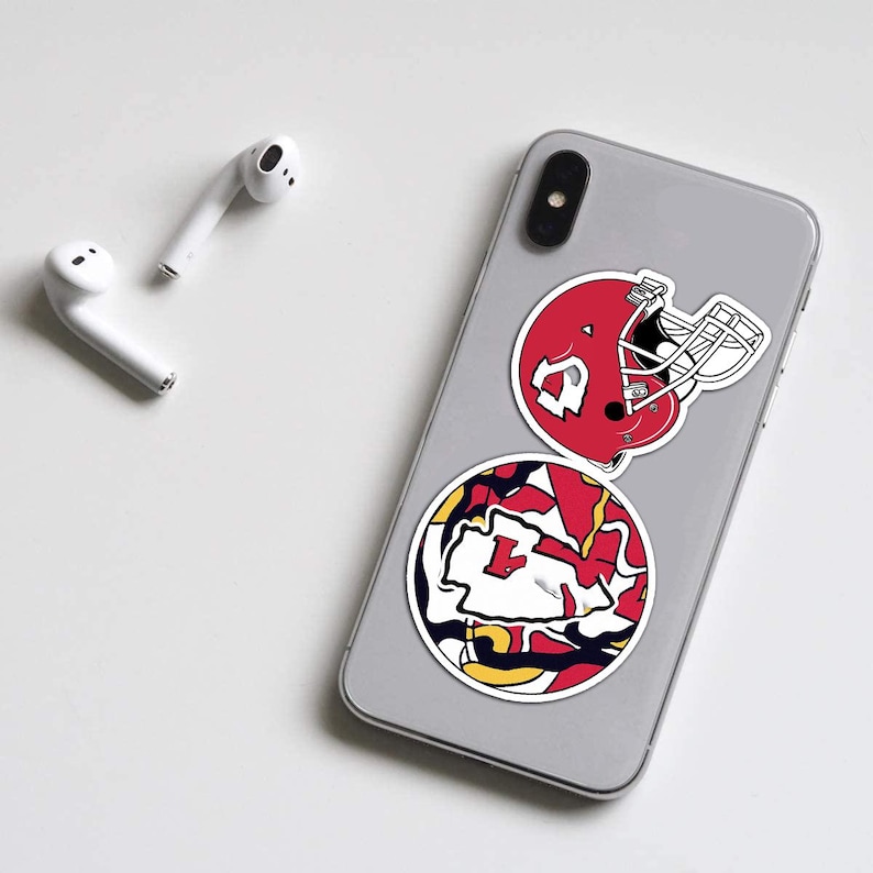 May include: A smartphone with two stickers featuring a red football helmet and a circular design with red, yellow, and white elements. White wireless earbuds are placed on the white surface.