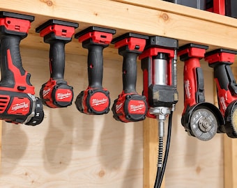 Può includere: Una collezione di sette utensili elettrici Milwaukee rossi e neri montati capovolti in fila sotto una mensola di legno. Gli utensili includono trapani, un ingrassatore e smerigliatrici angolari, tutti con il logo Milwaukee in bianco.