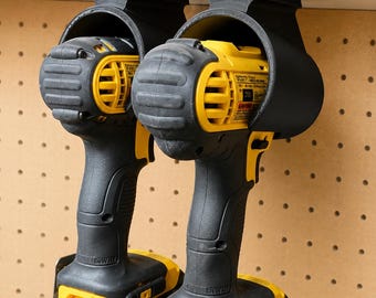 Puede incluir: Dos taladros inalámbricos negros y amarillos se guardan en soportes negros sujetos a una placa perforada. Los taladros tienen un cuerpo amarillo con detalles en negro y la marca "DEWALT" es visible.