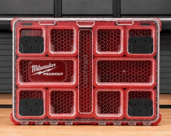 Può includere: Un organizer rosso Milwaukee Packout con coperchio trasparente dotato di molteplici scomparti interni. La custodia presenta un motivo a nido d'ape sui divisori interni e include inserti neri negli angoli. Il logo Milwaukee Packout è stampato sulla parte anteriore.
