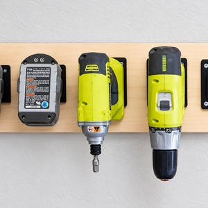 May include: A light-colored wooden wall mount organizer holding two gray battery packs, two lime green power tools, and one empty black mounting dock. The batteries feature white labels with black text and blue circular icons.
