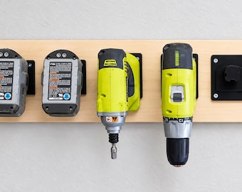 Può includere: Un supporto a parete in legno chiaro che sostiene due batterie grigie, due utensili elettrici verde lime e una base di montaggio nera vuota. Le batterie presentano etichette bianche con testo nero e icone circolari blu.