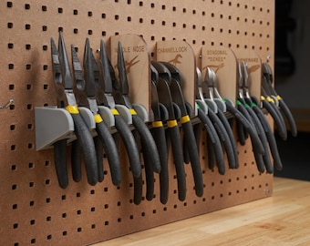 Può includere: Una collezione di pinze e tronchesi, tra cui pinze a becco e tronchesi diagonali, organizzata su una pannello forato. Gli utensili hanno manici neri con dettagli gialli e sono tenuti in supporti di plastica grigia. Etichette in legno identificano ogni tipo di utensile.