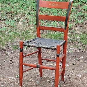 May include: A red wooden chair with a black woven seat and black painted accents. The chair has a simple design with four legs and a back with three horizontal slats.