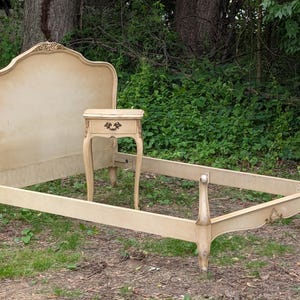 May include: A cream-colored wooden bed frame featuring a tall, arched headboard with carved floral detailing at the top, a matching footboard with curved posts, and a single-drawer nightstand with a dark metal pull handle and carved legs.