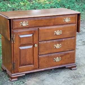 May include: A wooden buffet with a drop-leaf extension, four drawers, and a cabinet door with a raised panel design. The buffet has brass hardware and a dark wood finish.