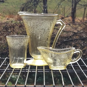 Puede incluir: Un juego de cristalería vintage de color amarillo, que incluye una jarra, un vaso y una crema. Las piezas presentan un diseño floral en relieve. La jarra tiene un asa geométrica. El juego se exhibe en una rejilla metálica negra.