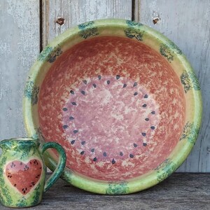 Puede incluir: Un juego de cuenco y jarra de cerámica con temática de sandía. El cuenco tiene un interior rojo con detalles de semillas negras y un borde verde. La jarra presenta un diseño de sandía en forma de corazón. El juego está pintado en tonos rojos, verdes y amarillos.