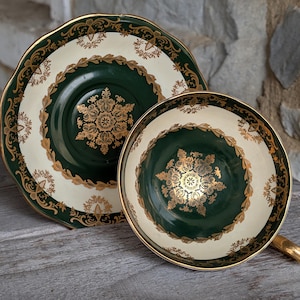 May include: A vintage teacup and saucer set. The teacup and saucer feature a cream-colored background with a dark green center and gold filigree detailing. The cup has a gold handle and rim, and the saucer has a scalloped edge.