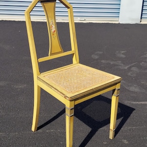 May include: A yellow painted chair with a woven seat and a decorative back. The back of the chair has a floral design. The chair has a rectangular seat and four legs. The chair is painted yellow with gold accents.