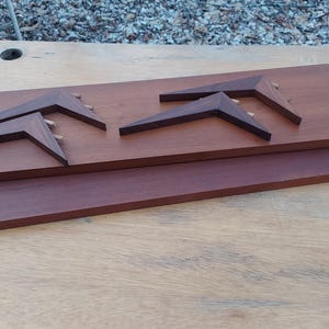 May include: A wooden wall art piece featuring three dark brown, angular shapes mounted on a rectangular, medium-brown wooden base. The base rests on a larger, darker brown wooden plank. The artwork is displayed on a light-colored wooden surface.