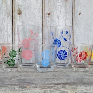 May include: A selection of clear drinking glasses featuring floral patterns in shades of pink, orange, green, blue, red, and yellow. The glasses are displayed on a wooden surface, set against a weathered wooden backdrop.