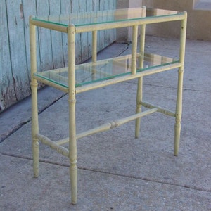 May include: A white painted metal shelf unit with two glass shelves. The shelf unit has a simple design with straight lines and rounded edges.