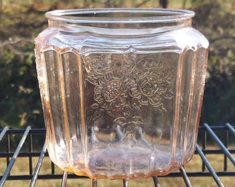 Pode incluir: Um frasco de vidro rosa vintage com um design floral. O frasco tem uma forma retangular com ranhuras verticais e uma abertura larga. O design floral é em relevo na frente. O frasco está sobre uma grelha de metal preta.