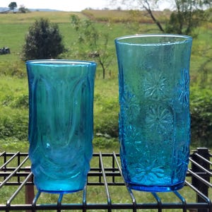 May include: Two vintage turquoise drinking glasses. The glass on the left has a textured, swirled design. The glass on the right features a floral pattern. Both glasses are made of thick glass and sit on a black metal rack.