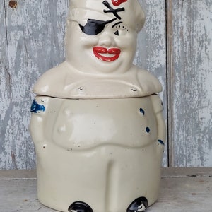 May include: A white ceramic cookie jar shaped like a cartoon pirate with a black eyepatch, red lips, and a blue and white sailor hat. The pirate is wearing a white shirt and blue trousers with black shoes.